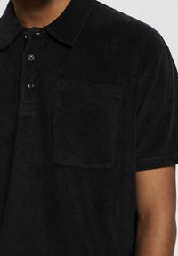 Gros plan sur un polo noir à manches courtes avec un tissu texturé, trois boutons et une poche poitrine sur le haut du corps d'une personne.