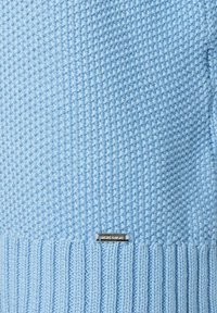 Helles blaues Strickpullover mit strukturiertem Muster, geripptem Saum und einem silbernen Logo-Tag am unteren Rand. Weicher, mitteldicker Stoff.
