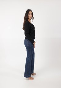 Cardigan negro acanalado con botones dorados, combinado con jeans de campana azul oscuro. El modelo lleva sandalias de tacón transparentes. Fondo de estudio.