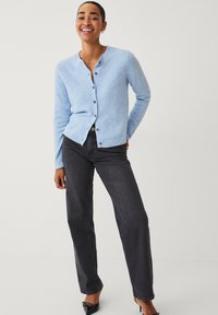 Cárdigan azul claro con cuello redondo y cierre de botones. Llevado con jeans oscuros y zapatos negros. Textura suave, diseño ajustado, mangas largas.
