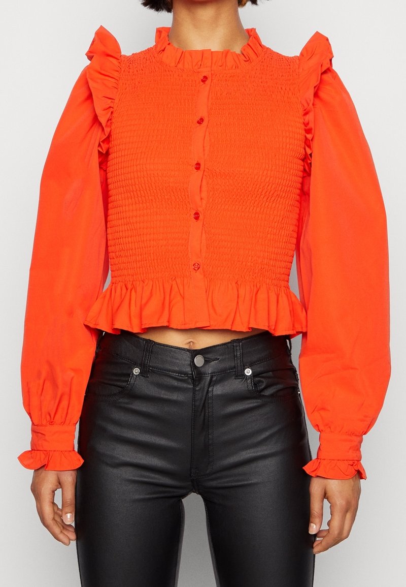 Blouse à manches longues orange vif avec col, poignets et ourlet à volants portée avec un pantalon en cuir noir taille haute.