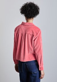 Camisa de pana coral con cuello, mangas largas y detalle de pliegue en la espalda, combinada con jeans azul oscuro. Patrón de tejido texturizado visible.