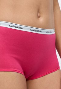 Růžové bavlněné kalhotky s bílým elastickým pasem, na kterém je černý nápis "Calvin Klein", s hladkou texturou a přiléhavým střihem.