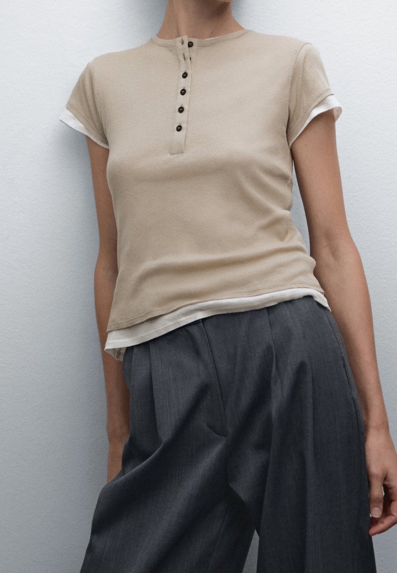 Haut beige à manches courtes avec un col rond, doté d'une patte de boutonnage et de poignets blancs contrastants, associé à un pantalon gris taille haute.