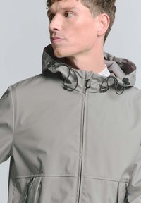 Lichtgrijze waterdichte jas met een capuchon, verstelbare trekkoorden en voorzakken met rits, met een zachte textuur.