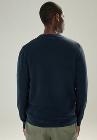 Mand vender væk iført en mørk marineblå langærmet sweater og lyse olivengrønne bukser mod en ensfarvet lys baggrund.