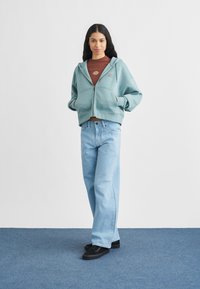 Νεαρή γυναίκα που στέκεται με τα χέρια στις τσέπες, φοράει μπλε φούτερ με φερμουάρ, καφέ μπλούζα, ανοιχτό μπλε τζιν και μαύρα αθλητικά παπούτσια.