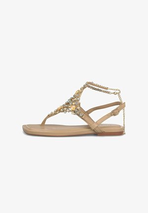 Sandalia plana beige con adornos de pedrería y gemas, tira en el tobillo con hebilla y cadena decorativa en el talón.