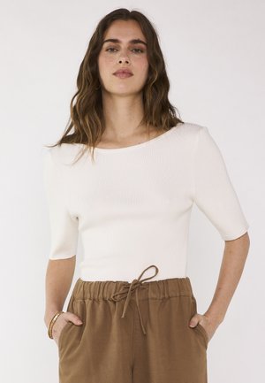 Donna con lunghi capelli mossi castani che indossa una maglietta bianca a maniche corte a coste e pantaloni marroni con coulisse, mani nelle tasche, sfondo neutro.