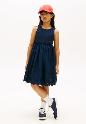 Ragazza che indossa un vestito senza maniche blu navy con orlo a occhielli, cappellino rosso, calzini bianchi e scarpe da ginnastica blu navy, in piedi con le gambe incrociate su sfondo bianco.