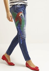Vaqueros de denim azul con un gran diseño de loro y hojas. Tienen un corte ajustado con detalles desgastados y se combinan con bailarinas rojas.