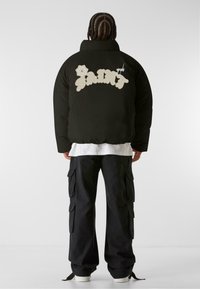 Giacca a puffo nera con una grafica crema "SAINT" e dettaglio floreale sulla parte posteriore. Indossata sopra una camicia bianca, abbinata a pantaloni cargo neri.