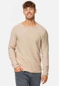 Beige pitkähihainen neule, jossa on teksturoitu neulekuviointi ja leveä kaula-aukko, yhdistettynä harmaisiin farkkuihin.