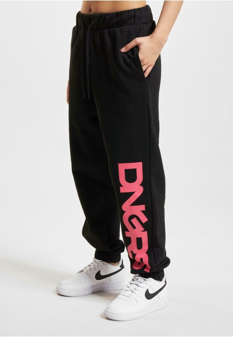 Crne trenirke s rozim "DNGRS" logotipom sa strane, elastična stražnja traka, bočni džepovi i rebrasti rubovi. Nosi se s bijelim Nike tenisicama.