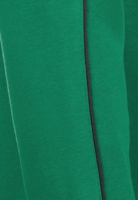 Tejido de sudadera verde con una superficie texturizada, que presenta un detalle de costura lateral negra a lo largo de toda la prenda. Costuras sutiles visibles.