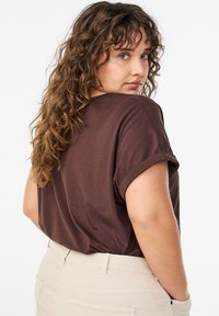 Dunkelbraunes T-Shirt mit aufgerollten Ärmeln, lockerer Passform, kombiniert mit hellbeigen Hosen. Weiche Textur, minimalistisches Design, keine sichtbaren Muster.