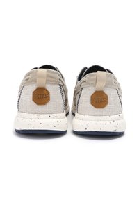 Beige fabric sneakers with a textured finish, hexagonal brown logo patch, and a speckled white sole; features neutral laces and back tab.