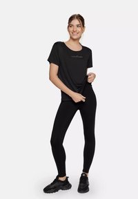 Schwarzes T-Shirt mit kurzen Ärmeln und Rundhalsausschnitt, bedruckt mit "CARDIO BUNNY" in Gold, kombiniert mit schwarzen Leggings und schwarzen Sneakers.