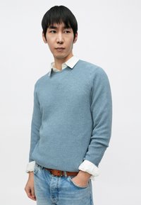 Svetlomodrý pletený sveter s textúrovaným povrchom, okrúhlym výstrihom a dlhými rukávmi, vrstvený cez bielu košeľu s golierom, v kombinácii s džínsami.
