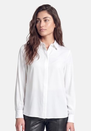 Woman with long wavy brown hair wearing a white button-up blouse and black leather pants, looking slightly to the side on a plain background.