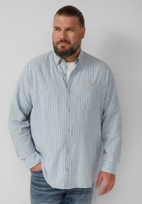 Knoop-overhemd met een blauw-wit geruit patroon, lange mouwen, een borstzak en tan knopen. Draagt over een wit T-shirt.