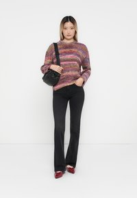 Femme portant un pull à rayures multicolores, un jean flare noir, des chaussures rouges et portant un sac à bandoulière noir sur un fond blanc uni.