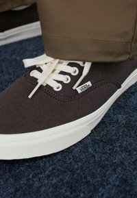 Brune lerrets-Vans-sneakers med hvite snørebånd og gummisåler. Viser logo og sømdetaljer, paret med kaki bukser mot en blå bakgrunn.