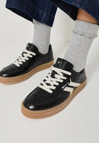 Černé nízké tenisky s texturou, bílé tkaničky a světle hnědé podrážky, nošené s světle šedými ponožkami s volánky a tmavě modrými kalhotami.