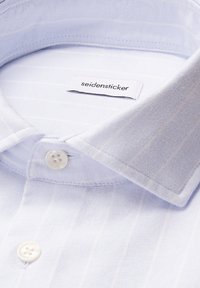 Primo piano di un colletto di una camicia a righe blu chiaro con bottoni bianchi e un'etichetta "Seidensticker" all'interno.