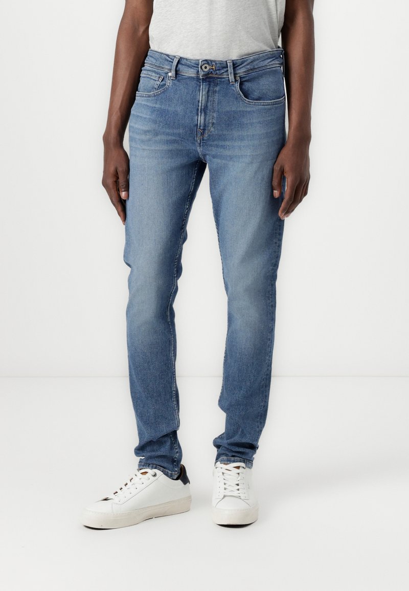 Jeans de mezclilla azul claro con un ajuste slim, cinco bolsillos y cierre de botón. Llevados con zapatillas blancas con acentos oscuros.
