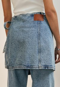 Svijetloplava traper suknja s neureznim rubom, koja ima kožnu oznaku sa natpisom "DENIM" i remen za struk. Tekstura tkanine s vidljivim šavovima.