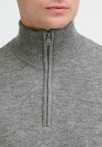 Harmaa neulepusero, jossa on korkea kaulus ja puolivetoketju. Siinä on pehmeä rakenne ja ribbikuviointi kauluksen ja vetoketjun lähellä.