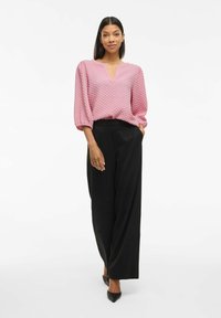 Kvinna iklädd en texturerad rosa blus med trekvartslånga ärmar och svarta vida byxor, stående med en hand i fickan och leende.