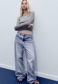 Grau langärmliges Shirt und hellblaue weit geschnittene Jeans. Der Denim hat eine ausgewaschene Textur mit sichtbaren Nähten und einem Fünf-Taschen-Design. Schwarze Sandalen.