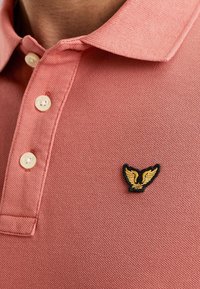 Svetloružová polo košeľa vyrobená z textúrovaného materiálu s tromi bielymi gombíkmi a čierno-zlatým krídlovým logom na ľavej hrudi.
