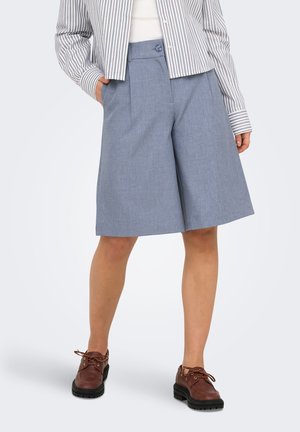 Personne portant un short gris large mi-genou, une chemise rayée à manches longues et des chaussures marron à lacets, debout les mains dans les poches.