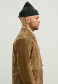 Mann in braunem Wildlederjacke und schwarzer Strickmütze, der nach rechts schaut, vor einem schlichten grauen Hintergrund.