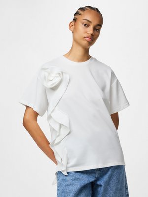 Personne portant un t-shirt blanc ample avec une grande fleur en tissu et un volant à l’épaule, assorti d’un jean bleu, debout devant un fond uni.