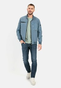 Blauwe bomberjack met een ritssluiting aan de voorkant, twee zakken en geribde manchetten. Gemaakt over een lichtgroen shirt en gecombineerd met donkere jeans en witte sneakers.