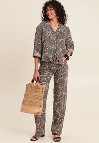 Twee-delig outfit met een loszittend, geknoopt shirt en bijpassende broek met een zwart-wit bladpatroon. Het model houdt een geweven beige tas vast.
