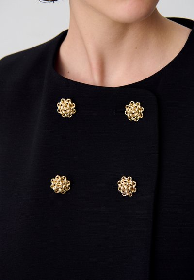Vêtement en tissu noir avec quatre boutons décoratifs en forme de fleur en or disposés en carré sur le devant.