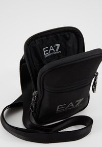Borsa a spalla in tessuto nero con chiusura a cerniera, dotata di un'etichetta interna. Include una lunga tracolla e una tasca con zip frontale.