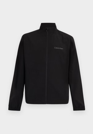 Fekete cipzáras dzseki magas gallérral, elasztikus mandzsettákkal és "Calvin Klein" logóval a bal mellrészen, egyszínű háttér előtt.