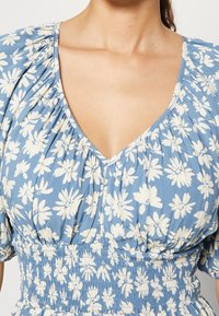 Blauwe bloemenjurk met een diepe V-hals, pofmouwen en een elastische taille. Dit model heeft witte bloemenpatronen op een zachte katoenen stof.