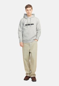 Sudadera gris con capucha ajustable, bolsillo frontal y gráfico negro. Combinada con pantalones sueltos de color claro y zapatos marrón oscuro.