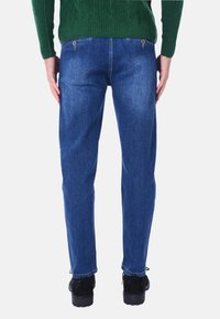 Jeans in denim blu con vestibilità dritta, dotati di una tasca posteriore con zip e una leggera sfumatura sbiadita, indossati con scarpe nere.