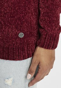 Bordeaux gebreide trui met geribbelde manchet en een zilveren logoknop. Draagt met lichtblauwe versleten jeans, met zichtbare handplaatsing.