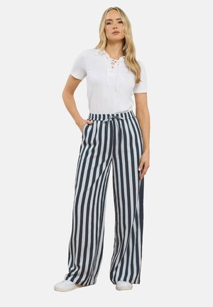 Kvinde iført hvid kortærmet snøreskjorte, høj taljede bukser med brede ben og sorte og hvide lodrette striber samt hvide sneakers, stående.