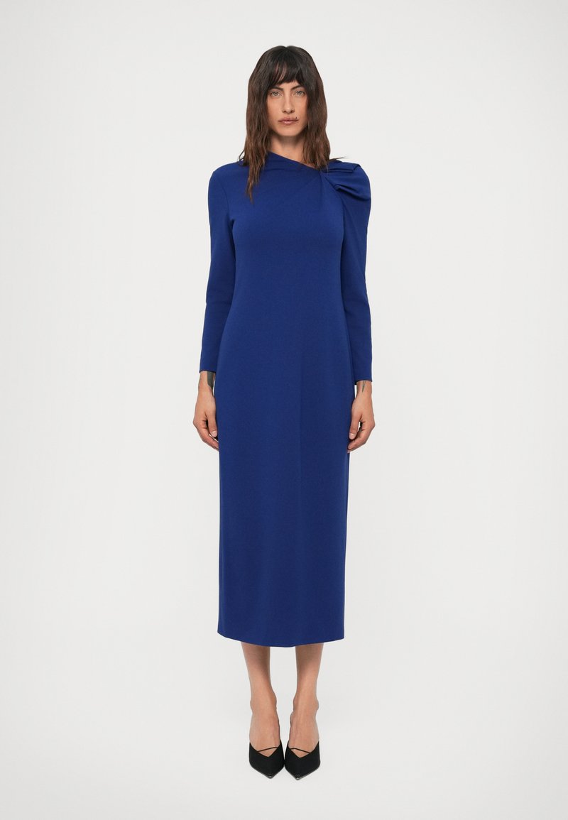 Mulher a usar um vestido midi de manga longa, azul royal, com um detalhe drapeado em um dos ombros, combinado com uns sapatos de salto preto de bico fino.