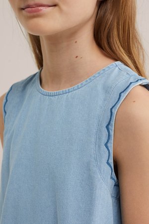 Fiatal személy, hosszú világos hajjal, ujjatlan világoskék felsőt visel, vállán fodros szélű részletekkel.